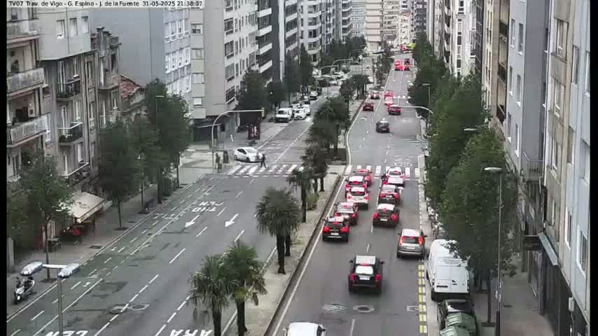 A city street with multiple cars driving in both directions, lined with buildings and trees, under a cloudy sky.