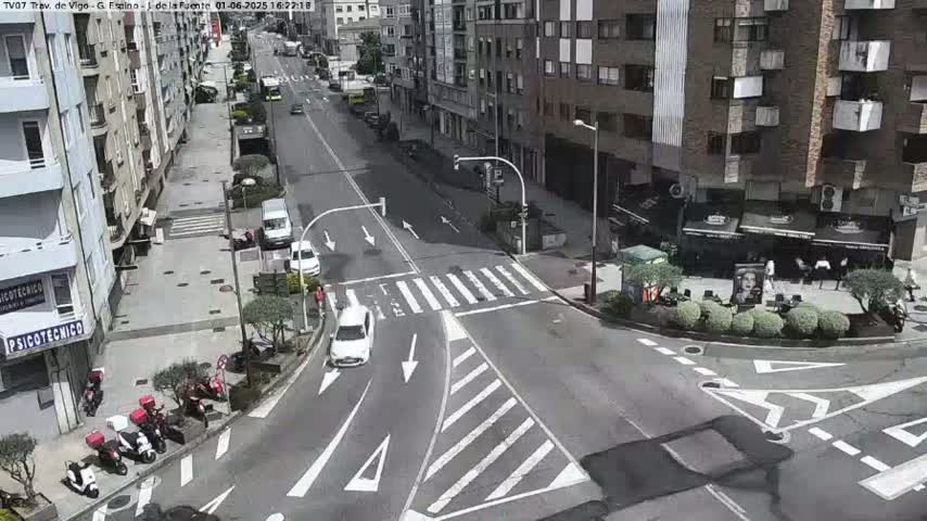 A sunny day reveals a city street intersection with several cars and parked motorcycles, bordered by multi-story buildings and outdoor seating areas.