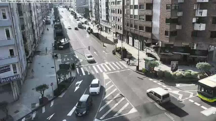 A sunny city street scene shows several cars at a crosswalk and intersection, with buildings lining the street and pedestrians walking on a sidewalk.