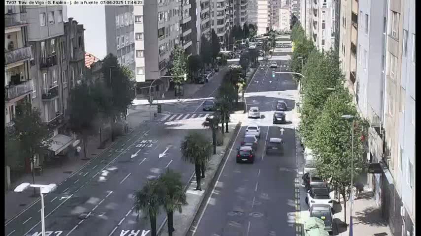 A city street scene on a sunny day shows several cars driving on a multi-lane road lined with trees and buildings.