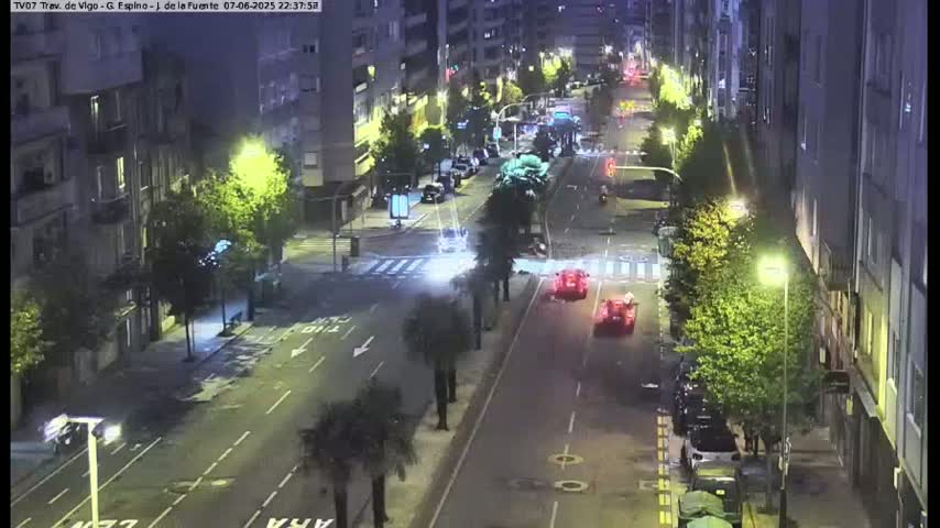 A nighttime, street-level view of a city street with cars driving on it, lined with buildings and trees, illuminated by streetlights.