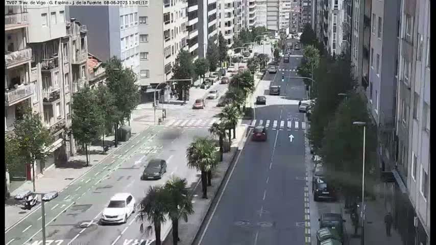 A sunny city street scene shows several cars driving on a multi-lane road lined with buildings and palm trees.