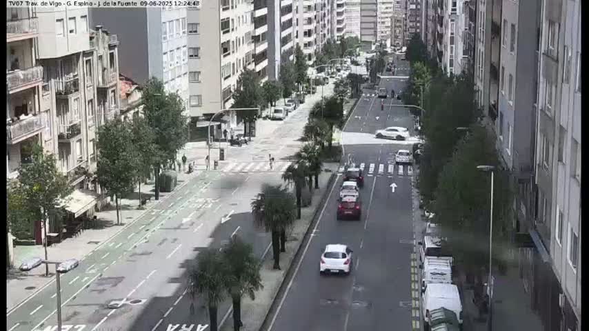 A city street scene on a sunny day shows several cars driving on a multi-lane road lined with palm trees and apartment buildings.