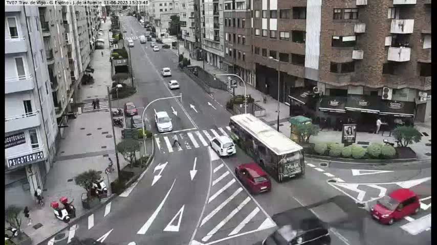A city street scene on an overcast day shows several cars and a bus at an intersection with pedestrians and parked motorcycles near buildings.