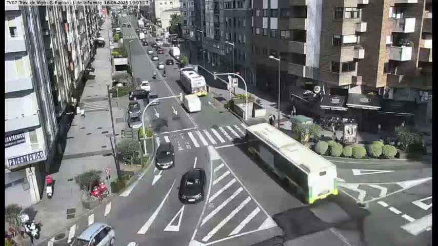 A sunny city street scene shows several cars and buses at a crossroad, with buildings lining both sides of the street.
