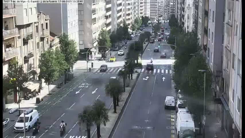 A city street scene on a sunny day shows several cars and vans driving on a multi-lane road, with tall buildings lining both sides, and palm trees planted along a median.