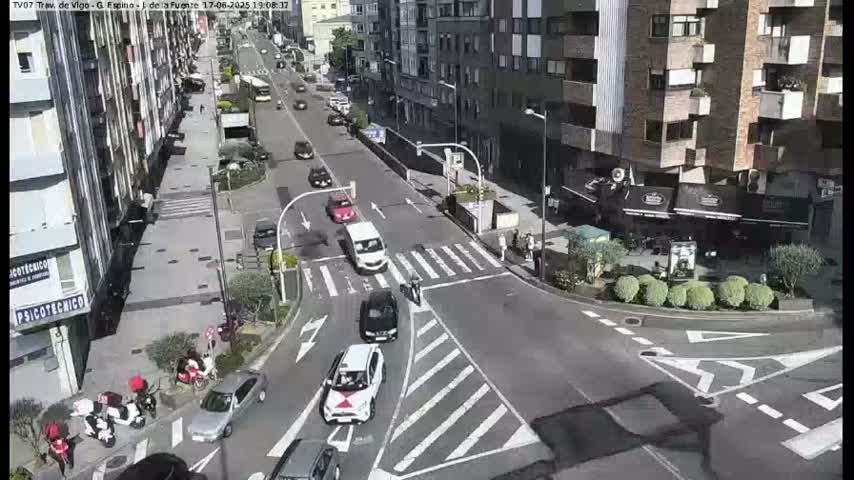 A sunny city street intersection shows several cars stopped at crosswalks and traffic signals, with pedestrians and delivery scooters on the sidewalk.