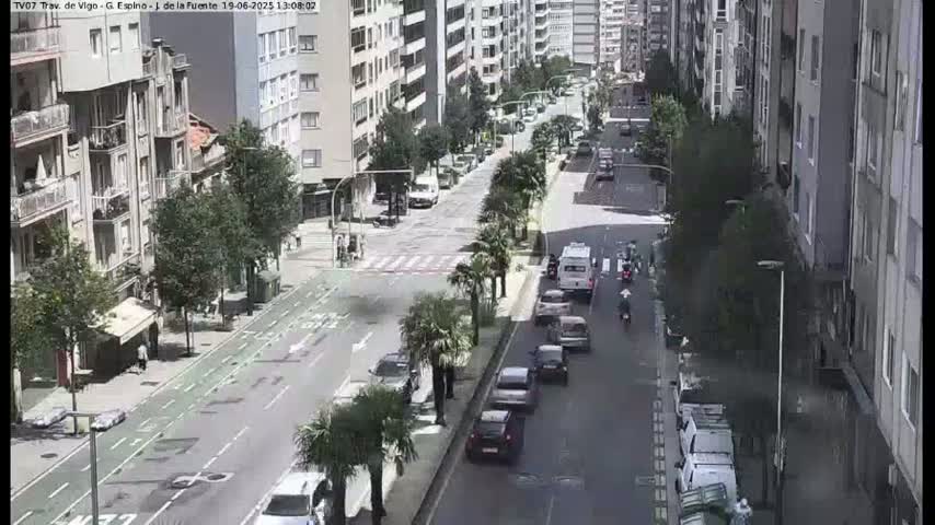 A city street scene on a sunny day shows several cars and pedestrians moving along a road lined with trees and multi-story buildings.