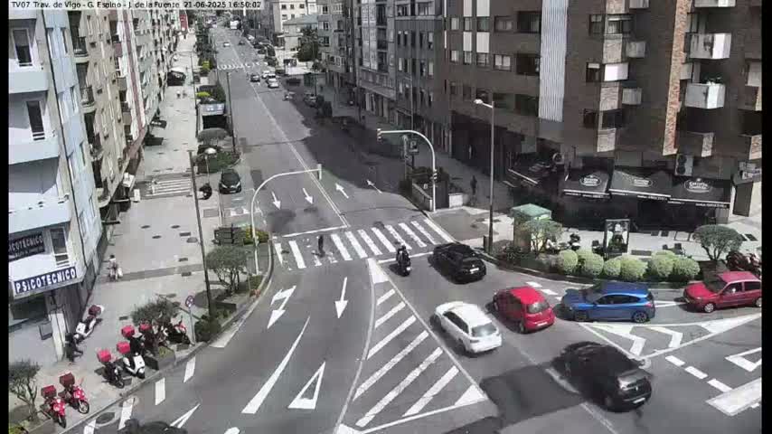 A sunny city street scene shows several cars at a crosswalk intersection, with pedestrians and motorcyclists also present, near several buildings and parked mopeds.
