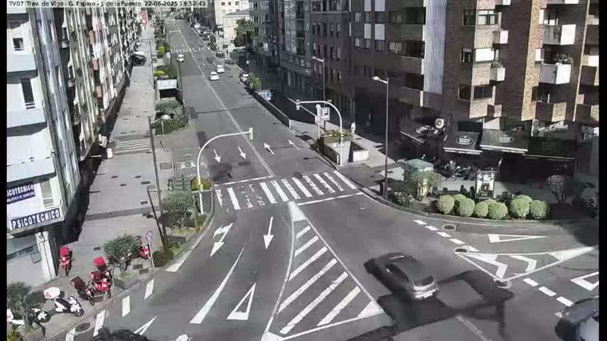 A sunny city street intersection shows several cars and delivery scooters, with pedestrians and buildings visible in the background.