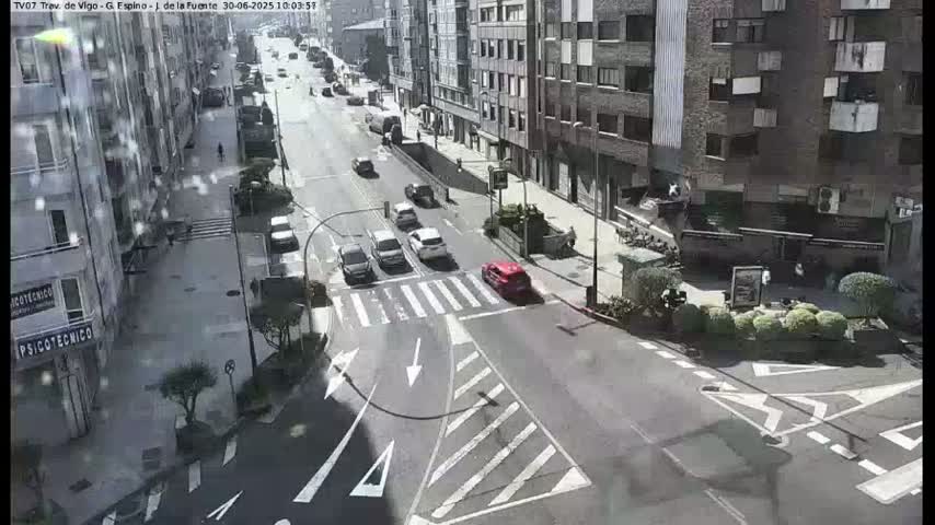A sunny city street scene shows several cars driving on a multi-lane road with pedestrian crosswalks, alongside buildings.