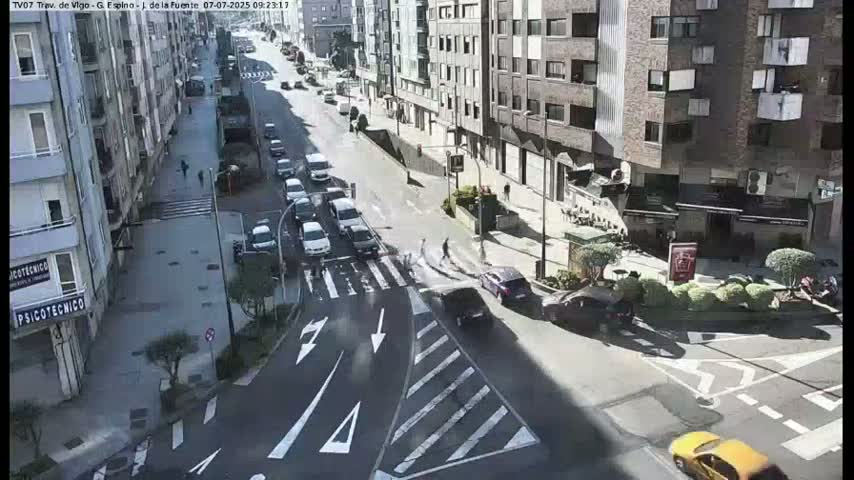 A sunny city street scene shows several cars driving on a roadway intersecting with a crosswalk, and buildings line both sides of the street.