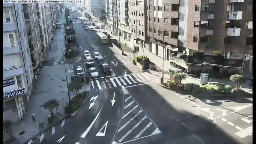 A sunny city street intersection shows several cars in motion and parked along the sides of buildings.