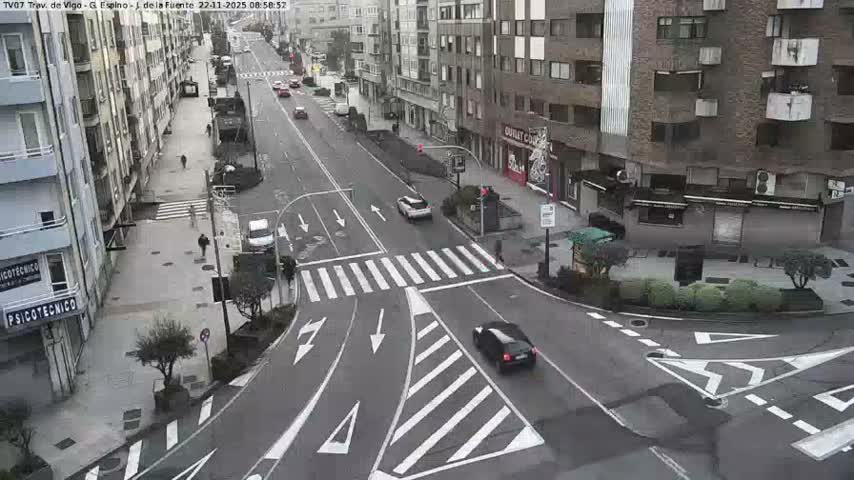 A damp, overcast urban street intersection is visible, bustling with a few cars and pedestrians navigating the road and sidewalks lined by multi-story buildings.