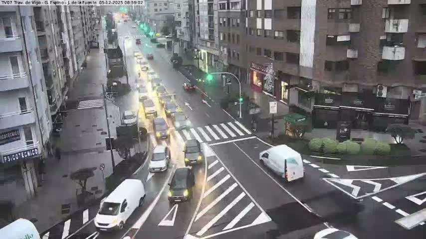 A busy urban street is seen from an elevated perspective, with numerous vehicles traveling along a wet road lined by multi-story buildings, under overcast conditions suggesting recent or ongoing rain.