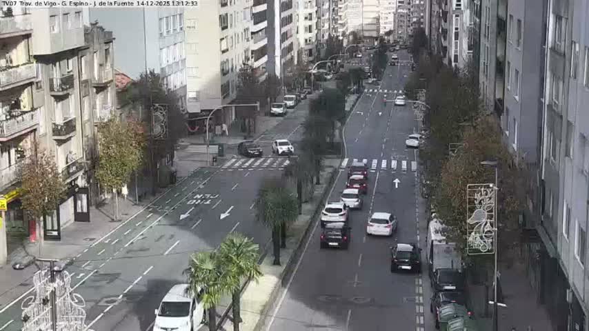 A busy urban street is seen from an elevated perspective, with numerous vehicles traveling along a wet road lined by multi-story buildings, under overcast conditions suggesting recent or ongoing rain.