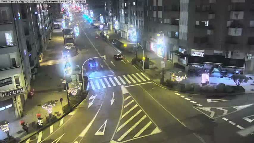 A busy urban street is seen from an elevated perspective, with numerous vehicles traveling along a wet road lined by multi-story buildings, under overcast conditions suggesting recent or ongoing rain.