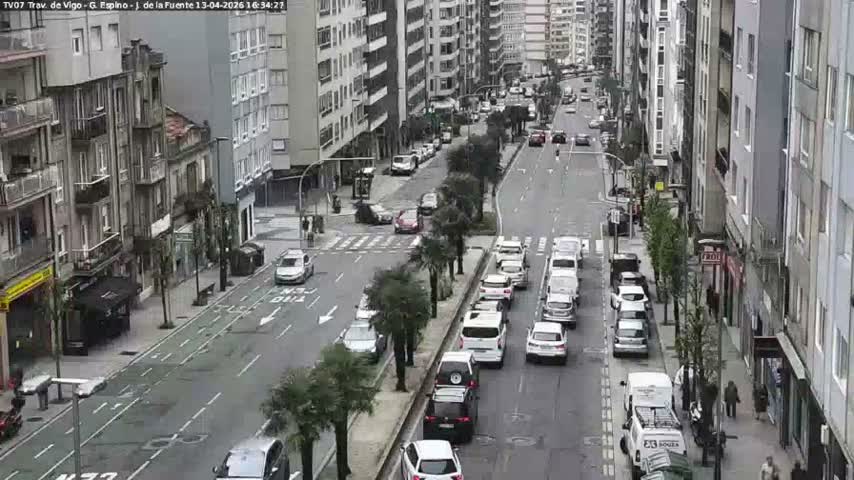 A busy urban street is seen from an elevated perspective, with numerous vehicles traveling along a wet road lined by multi-story buildings, under overcast conditions suggesting recent or ongoing rain.