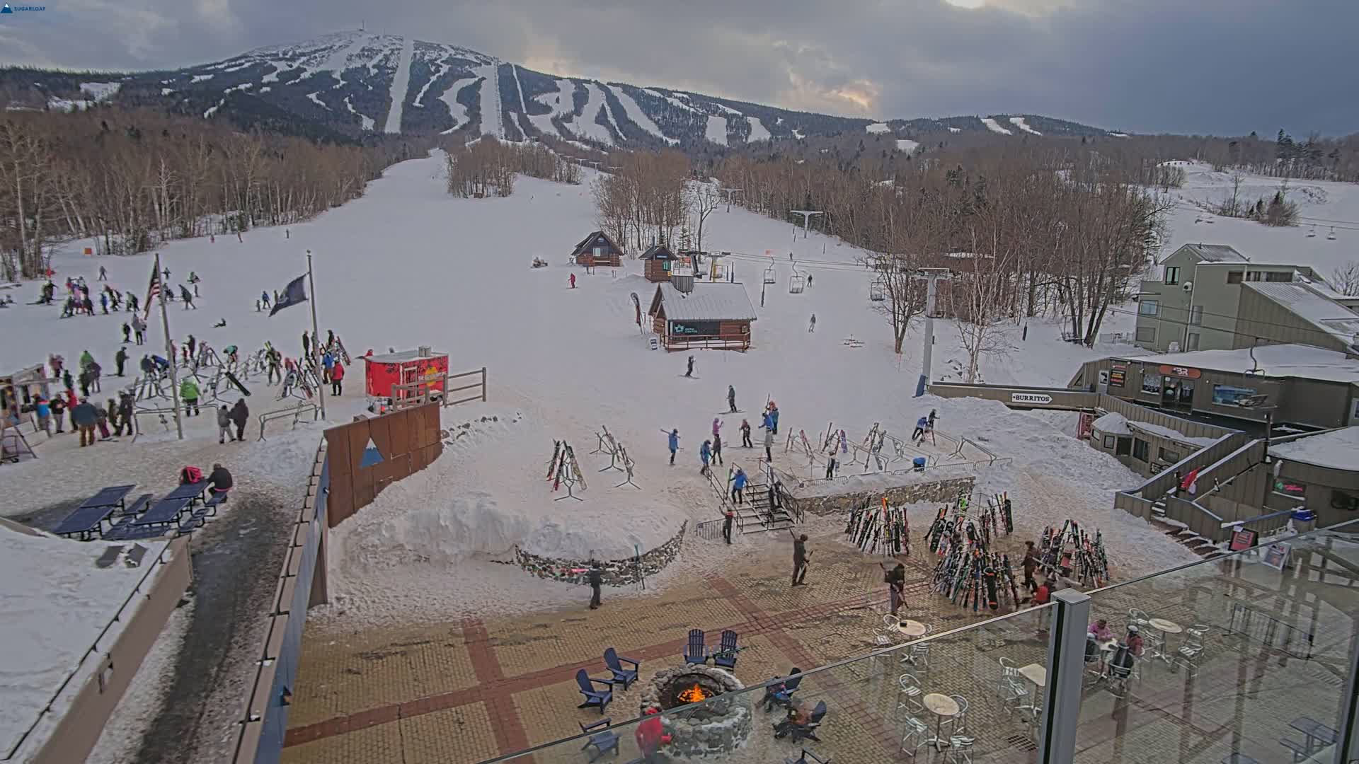 A snowy ski resort base area with people gathered around a fire pit, skiers on the slopes, and numerous skis leaning against racks, all under a cloudy sky.