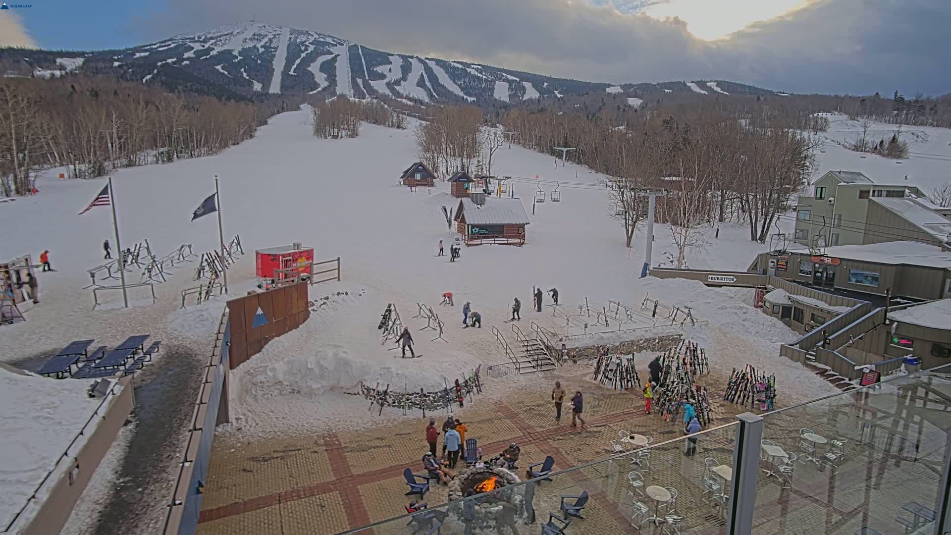 A snowy ski resort base area with a fire pit surrounded by people and ski equipment, situated below a mountain with ski runs under cloudy skies.