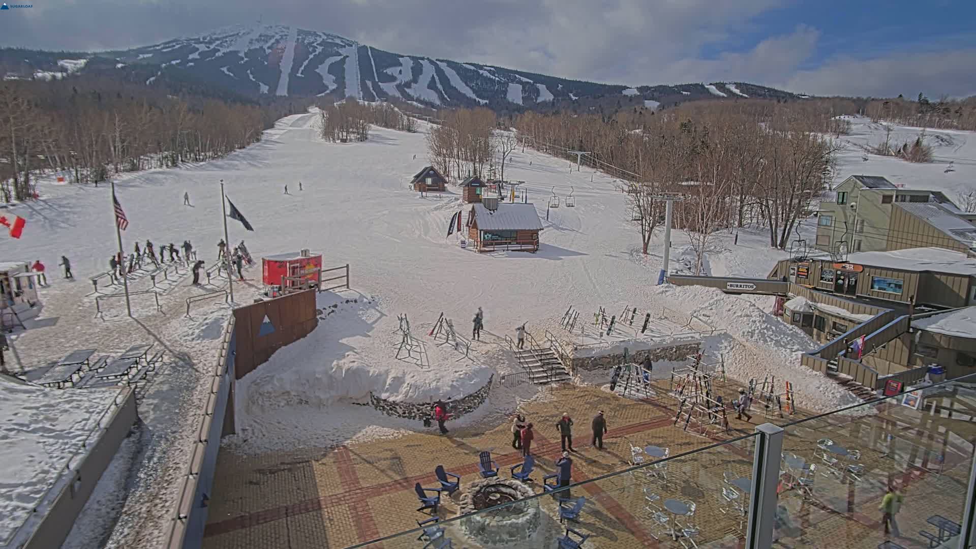 A sunny day at a snow-covered ski resort shows a base area with people, ski equipment, and buildings, with a mountain in the background.