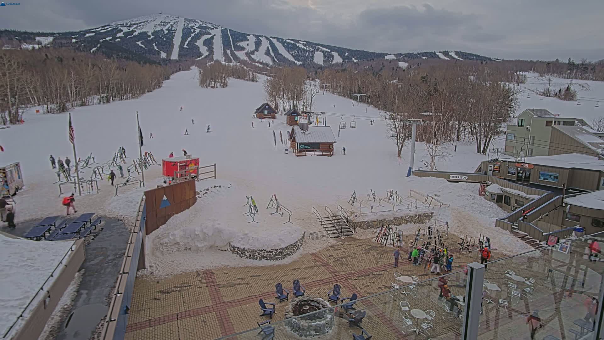 A snowy ski resort with several buildings, skiers, and a fire pit is visible under an overcast sky.