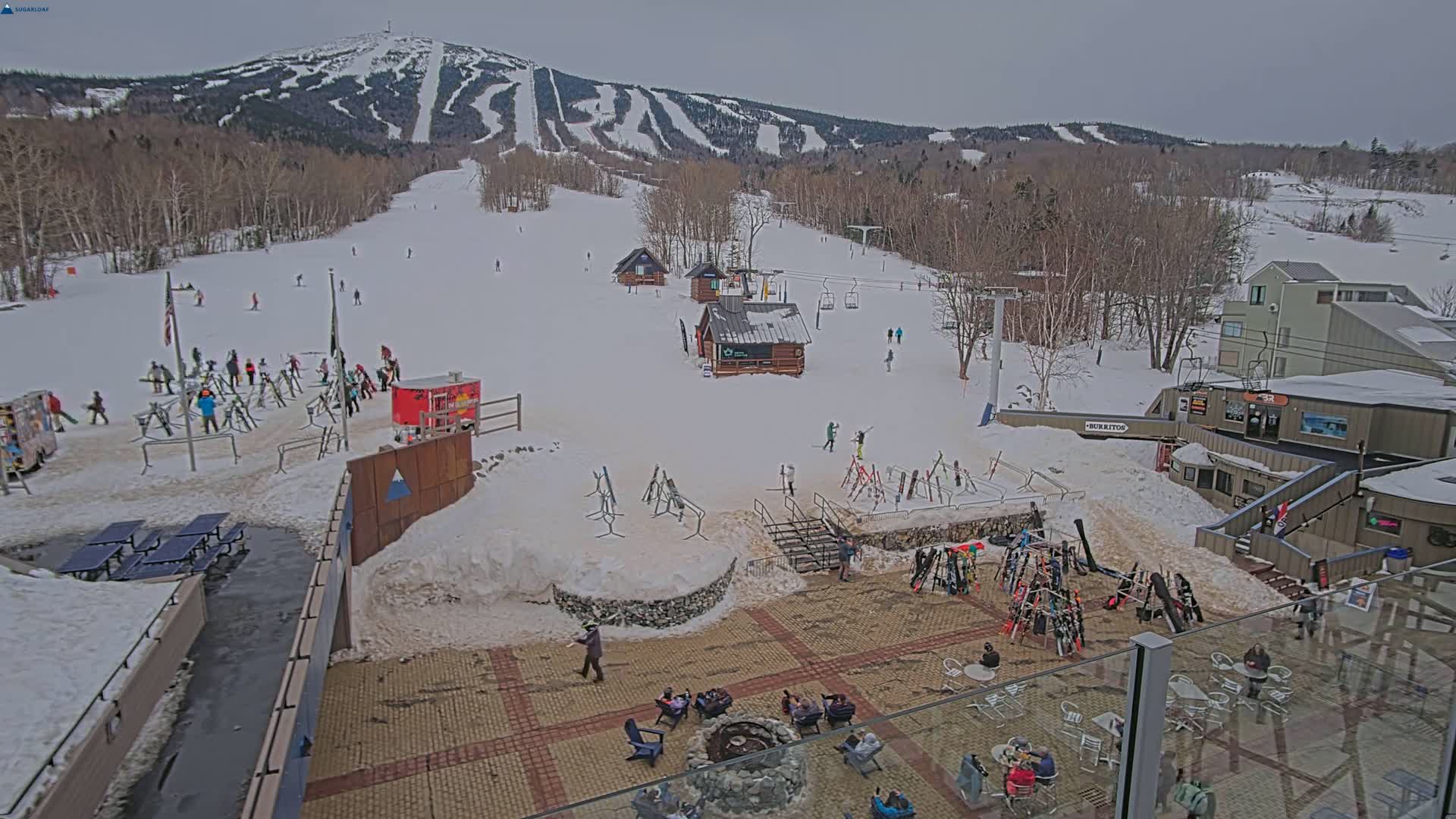 A snow-covered ski resort with people skiing and relaxing in an outdoor patio area on an overcast day.