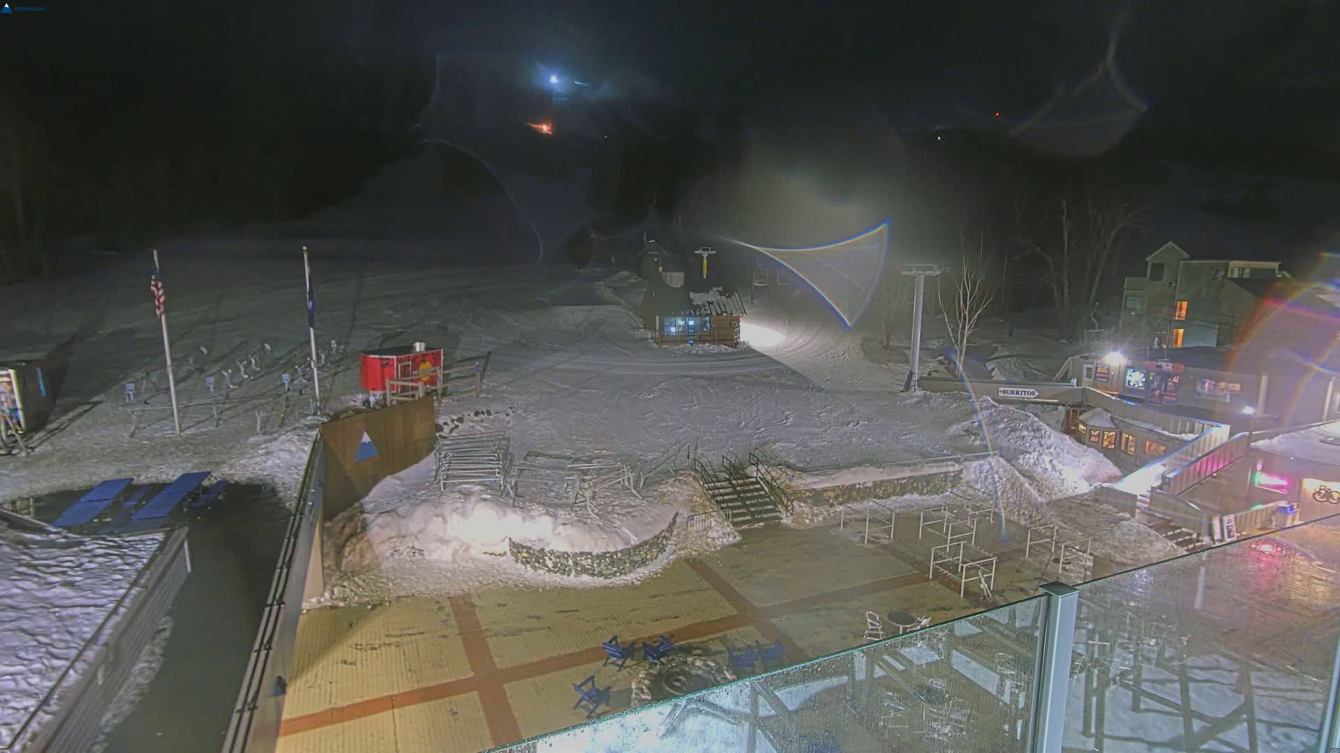 A snow-covered nighttime ski resort base area with a ski lift, buildings, and outdoor seating.