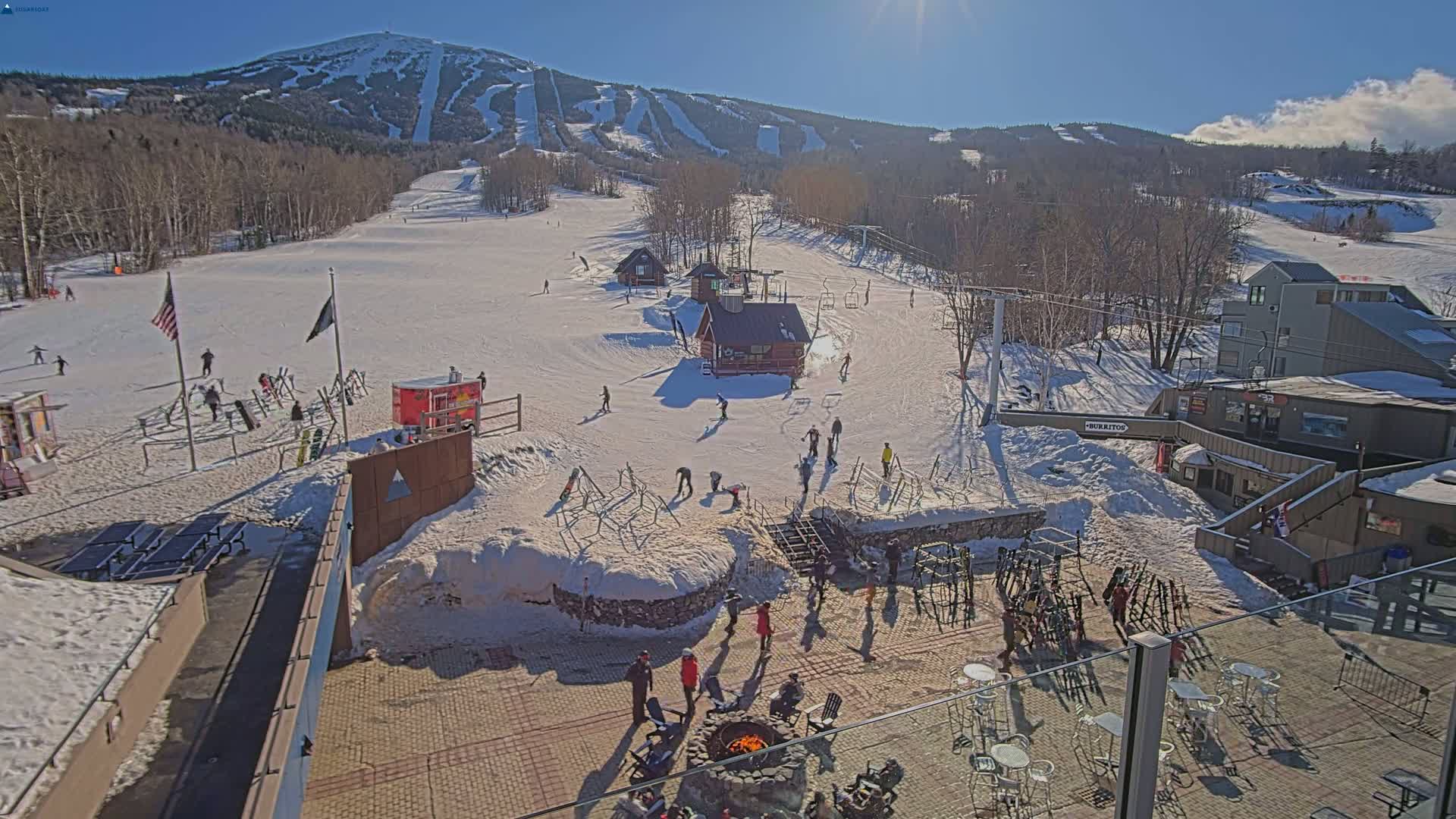 A sunny day at a snow-covered ski resort shows skiers and snowboarders on the slopes, with several small buildings and a fire pit in the foreground.