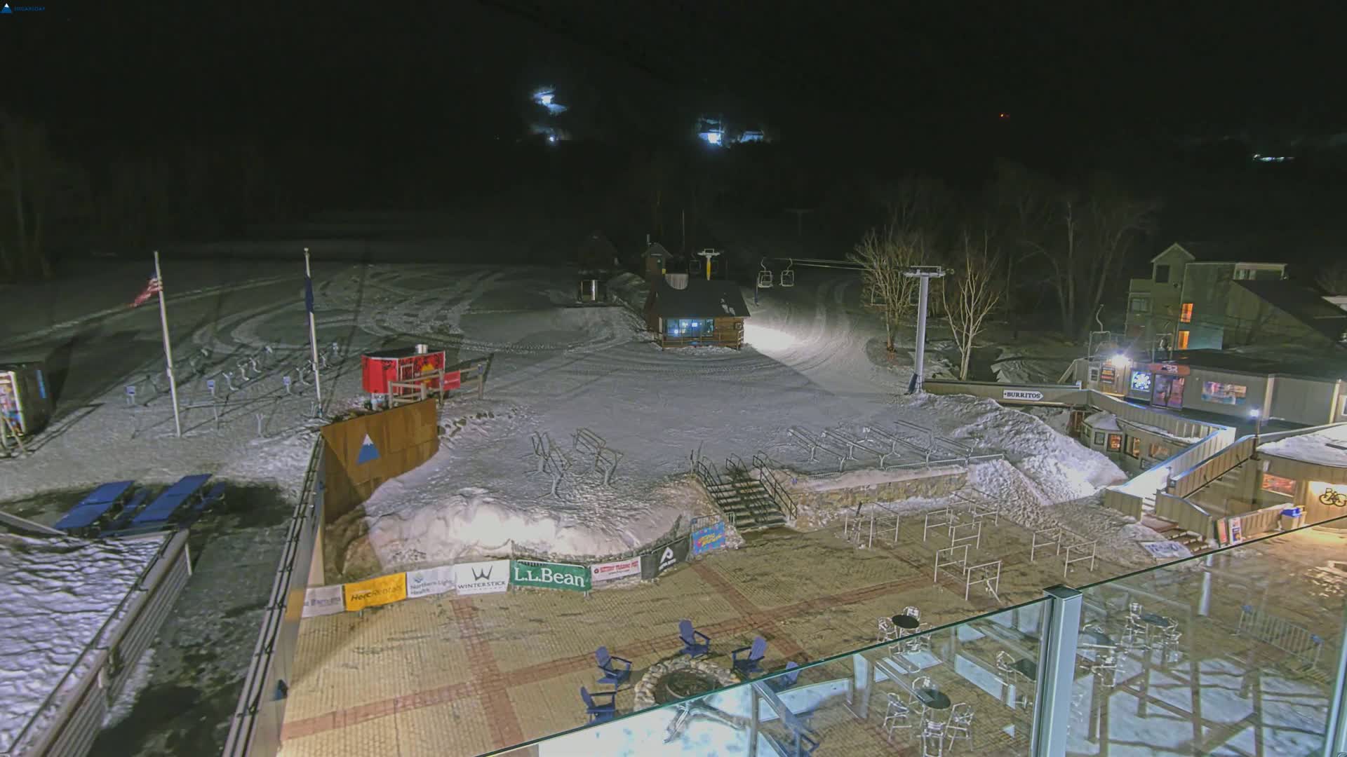 A snow-covered ski resort at night, illuminated by artificial lights, with a ski lift and several buildings visible.