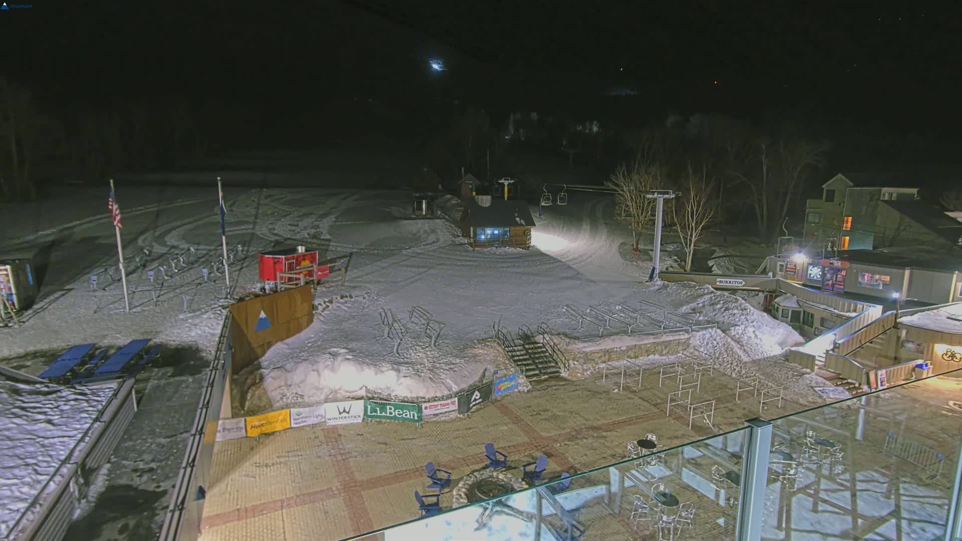 A nighttime view of a snow-covered ski area with a chairlift, a small building, and outdoor seating areas illuminated under a dark sky.