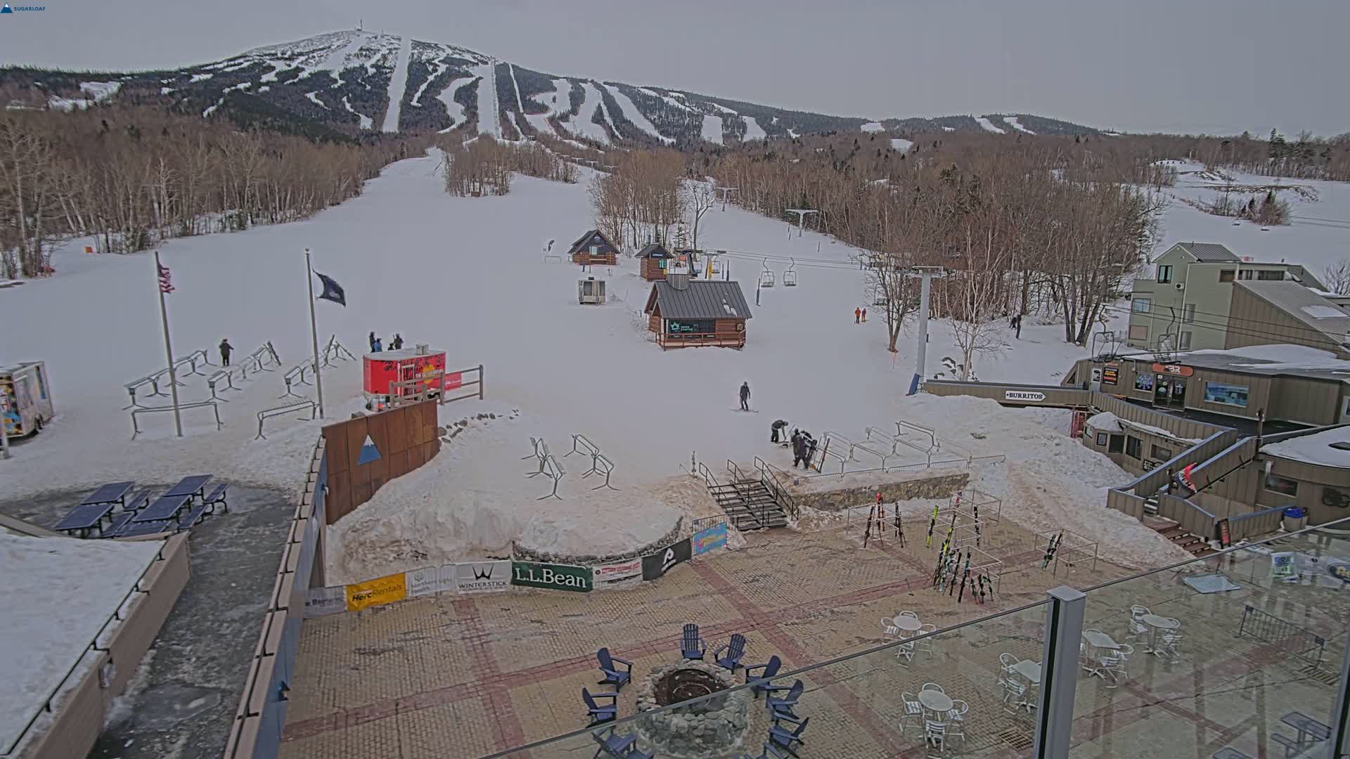 A snow-covered ski resort base area with several buildings, ski lifts, and people, under an overcast sky.