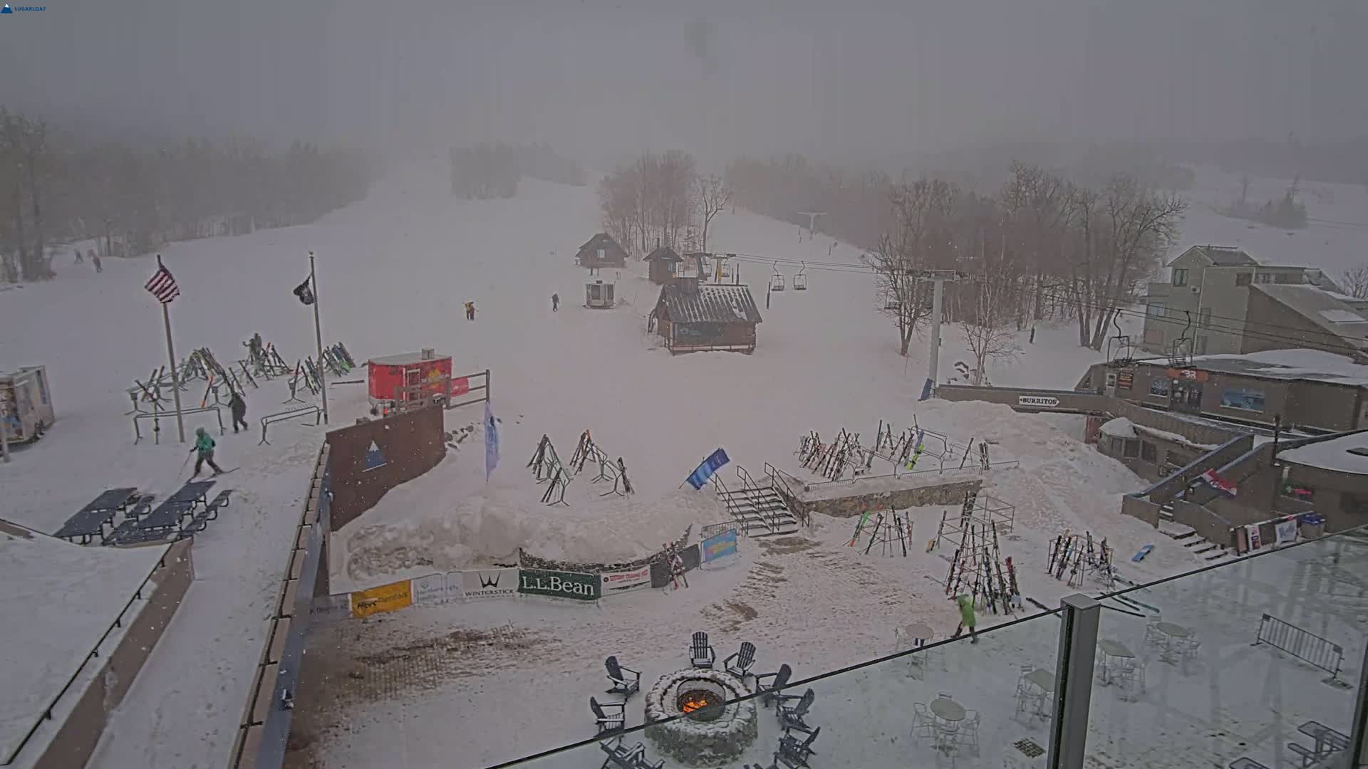 A snowy ski resort is seen on an overcast day, with light snowfall, featuring several buildings, ski lifts, and skiers scattered across the snow-covered slopes.