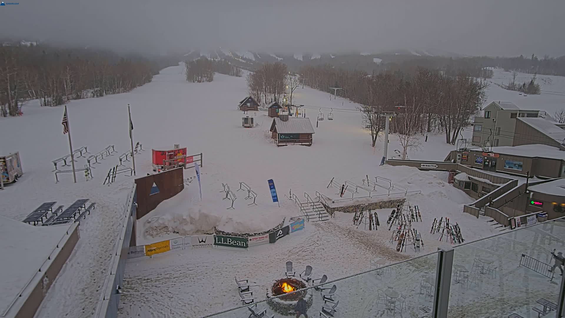 A snow-covered ski resort with several buildings, ski lifts, and outdoor seating areas is visible under an overcast sky.