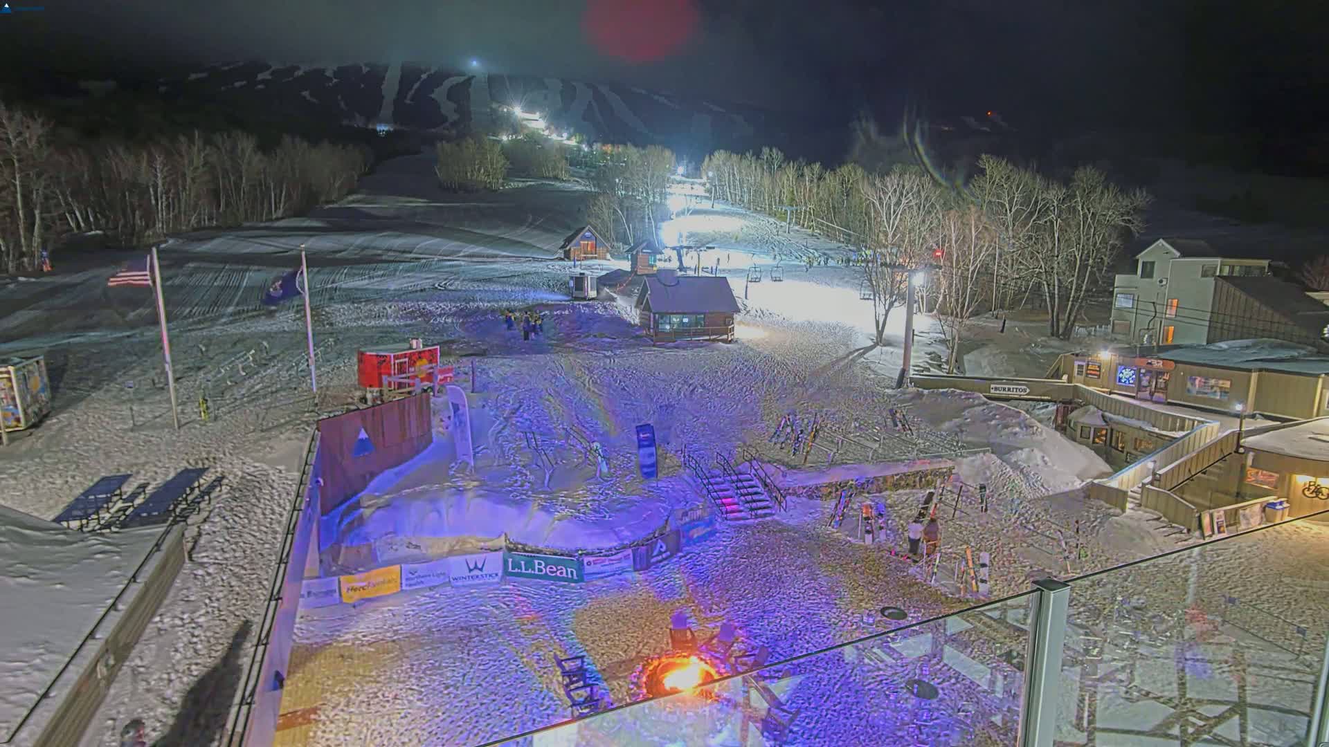 A nighttime view of a snow-covered ski resort with illuminated slopes, buildings, and a bonfire in the base area.