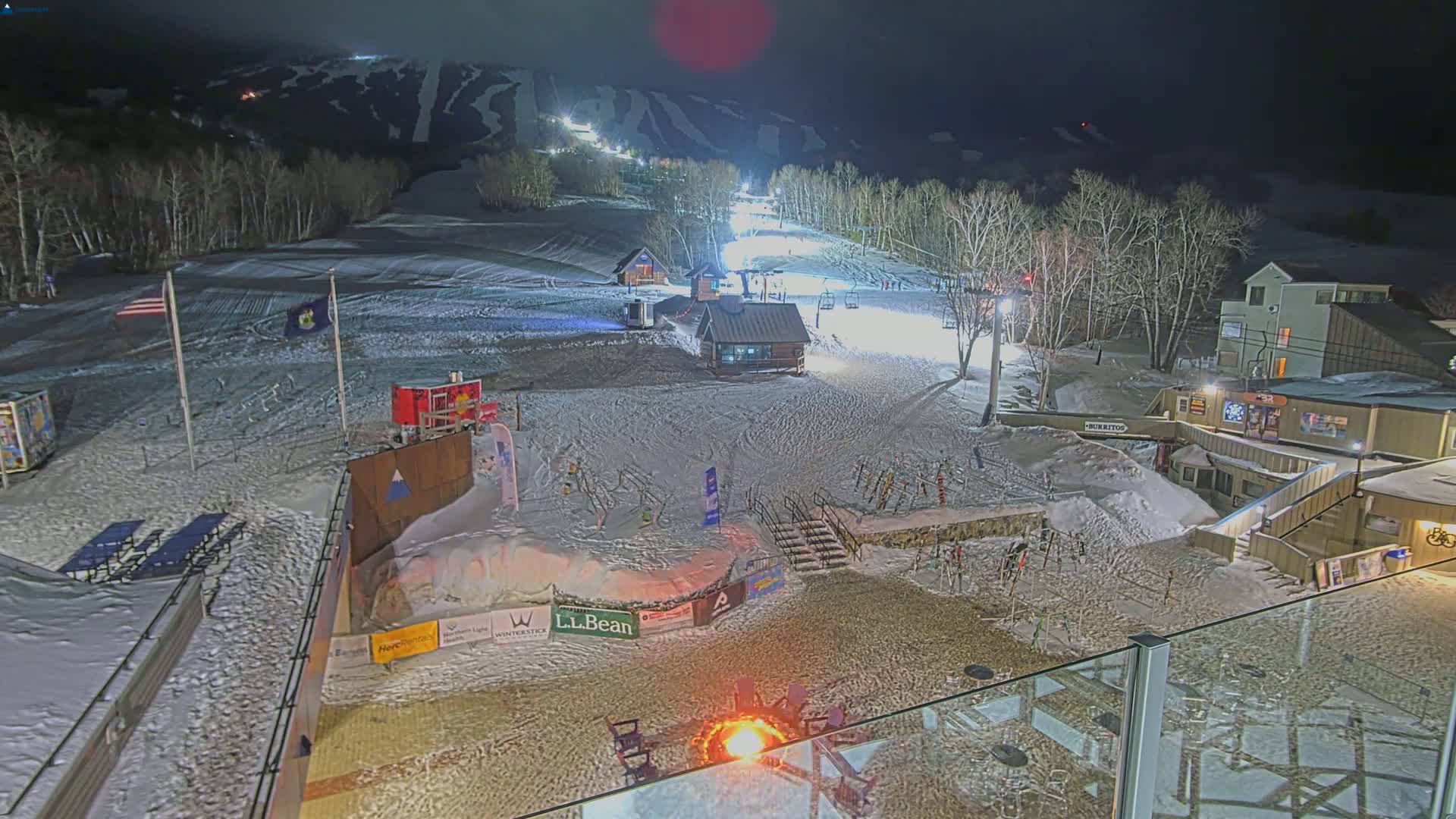 A snow-covered ski resort at night, illuminated by lights on the slopes, with a fire burning in the foreground.