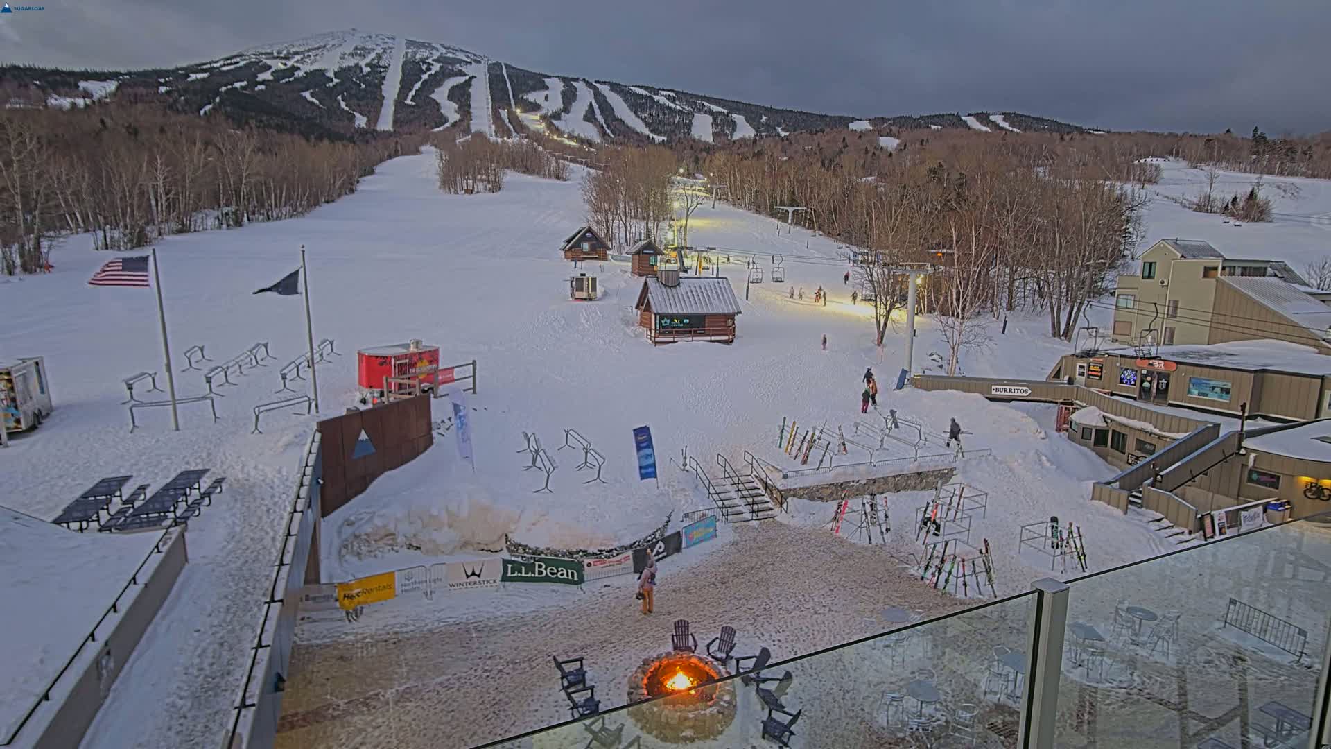 A snow-covered ski resort at dusk, with skiers and snowboarders visible on the slopes and in the base area, a fire pit burning in the foreground, and a mountain range in the background.