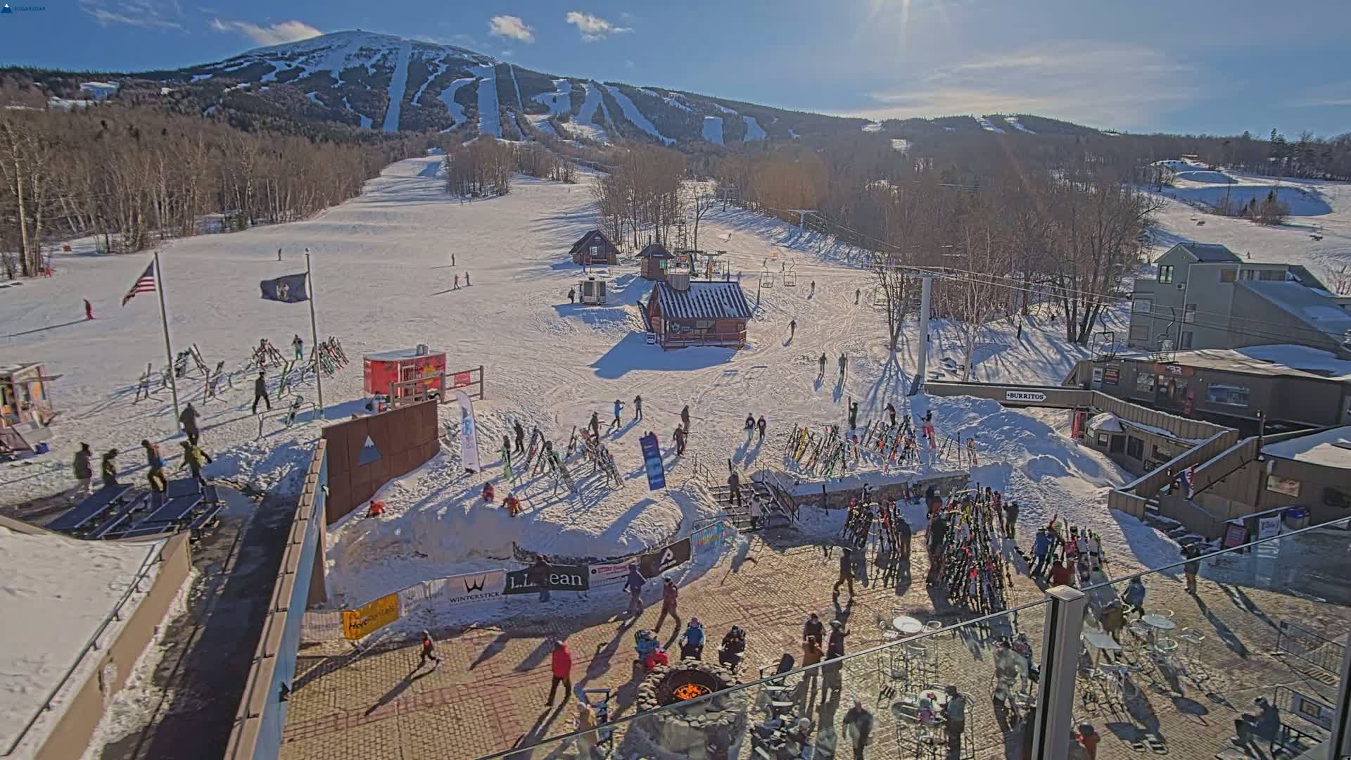 A sunny winter day at a ski resort shows skiers and snowboarders on the slopes and gathered in a plaza at the base of a snow-covered mountain.