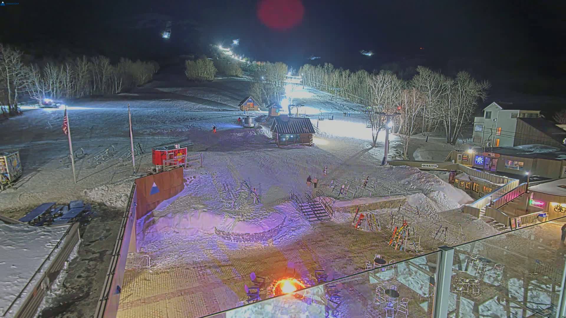 A nighttime view of a snow-covered ski slope with several buildings, a bonfire, and skiers visible under the lights.