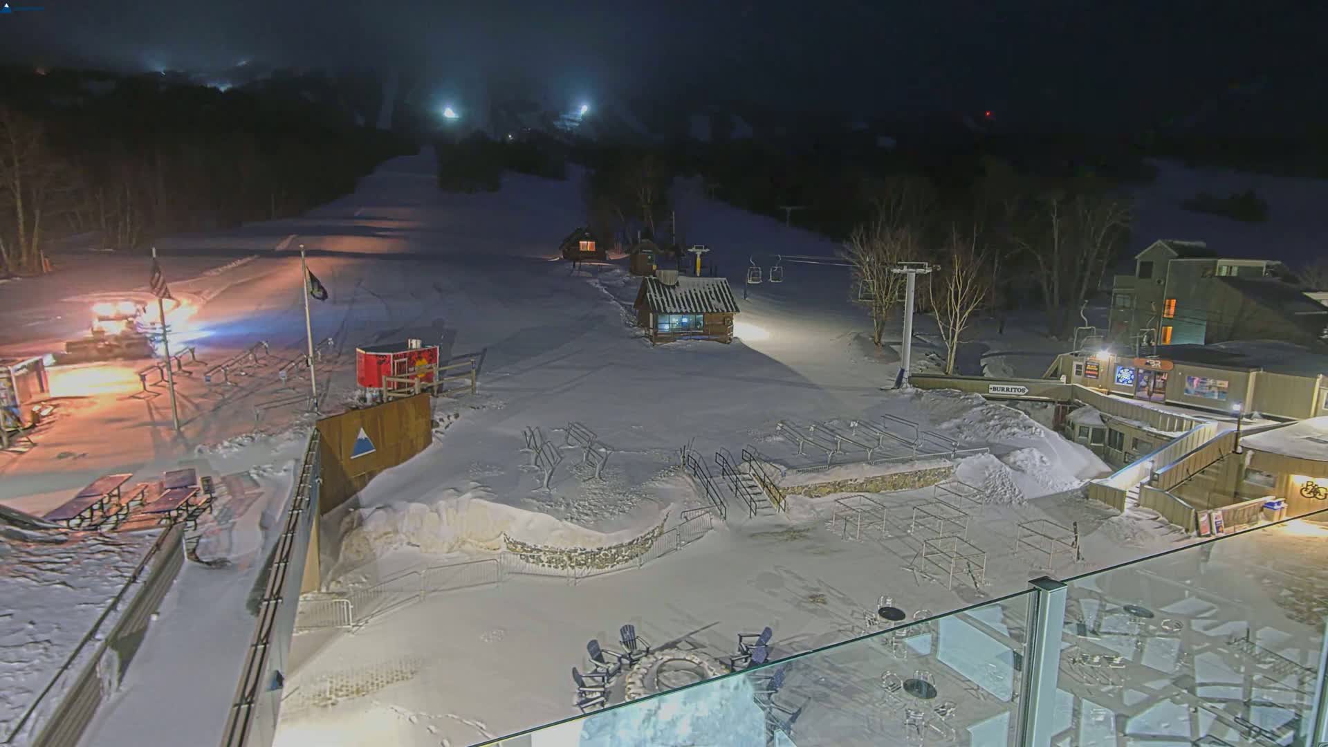 A snow-covered ski resort at night, illuminated by artificial lights, features a snowcat, ski lift, small buildings, and outdoor seating areas.