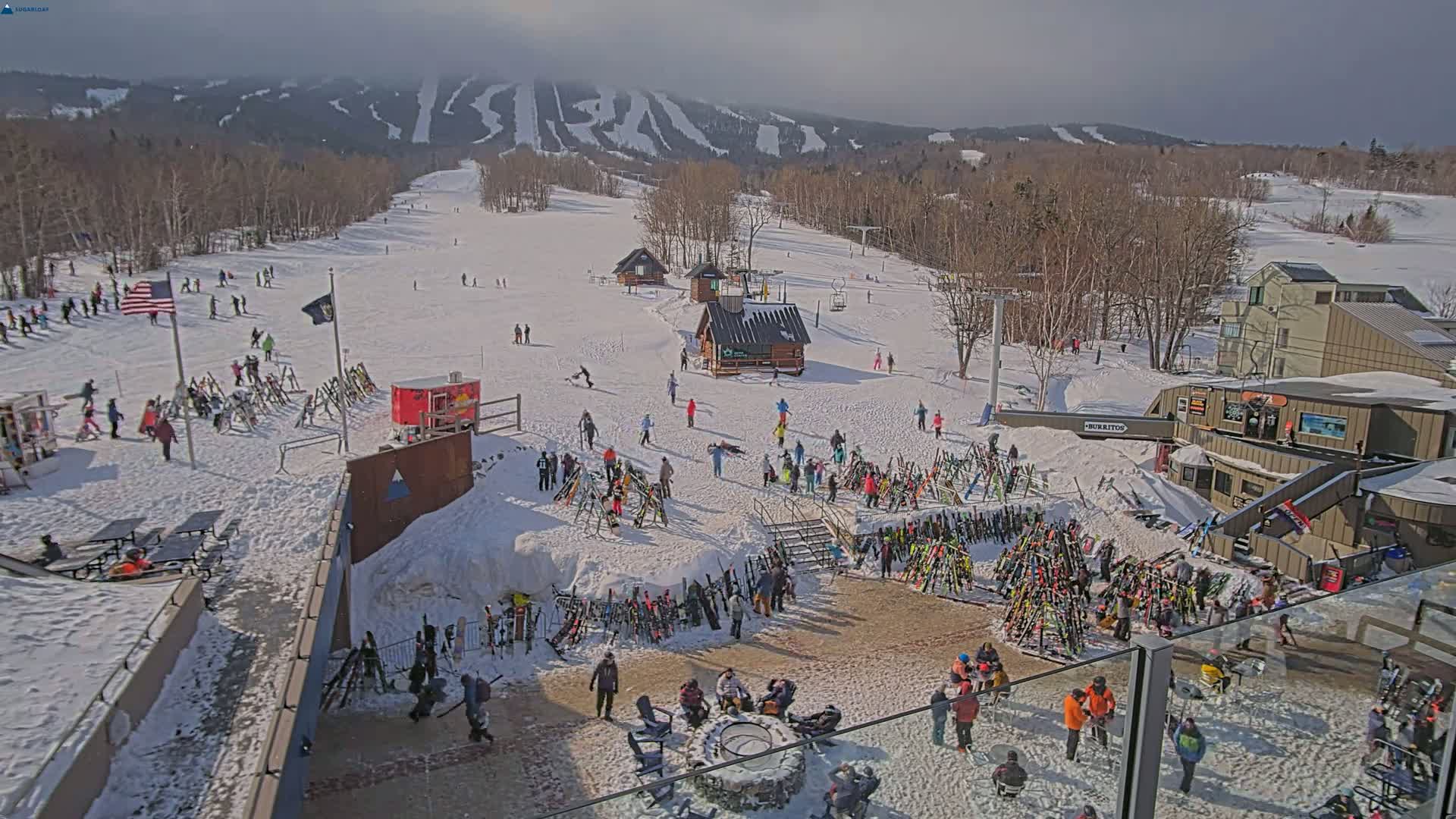 A snowy ski resort with numerous skiers and snowboarders, ski racks, and buildings on a sunny day with a mountain range in the background.