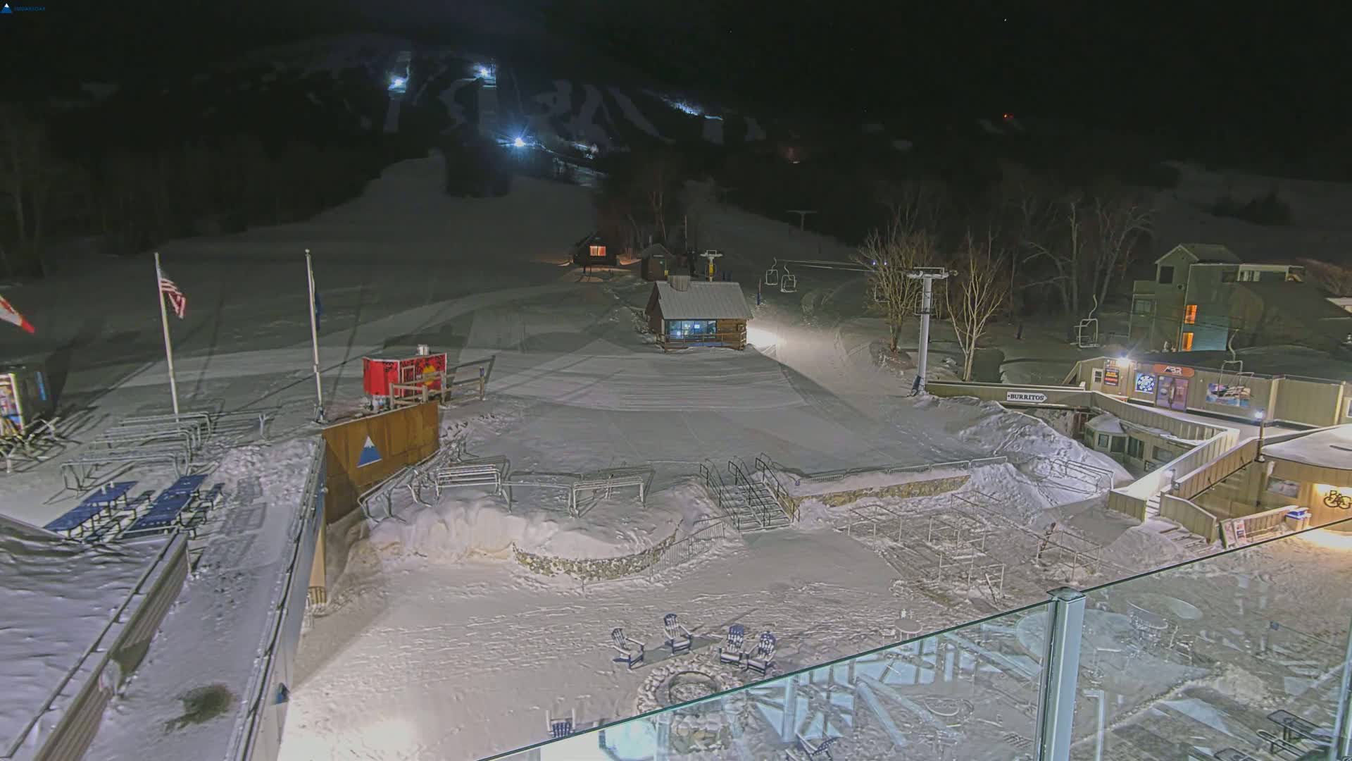 A snow-covered ski resort at night, illuminated by artificial lights, features ski lifts, buildings, and outdoor seating areas.