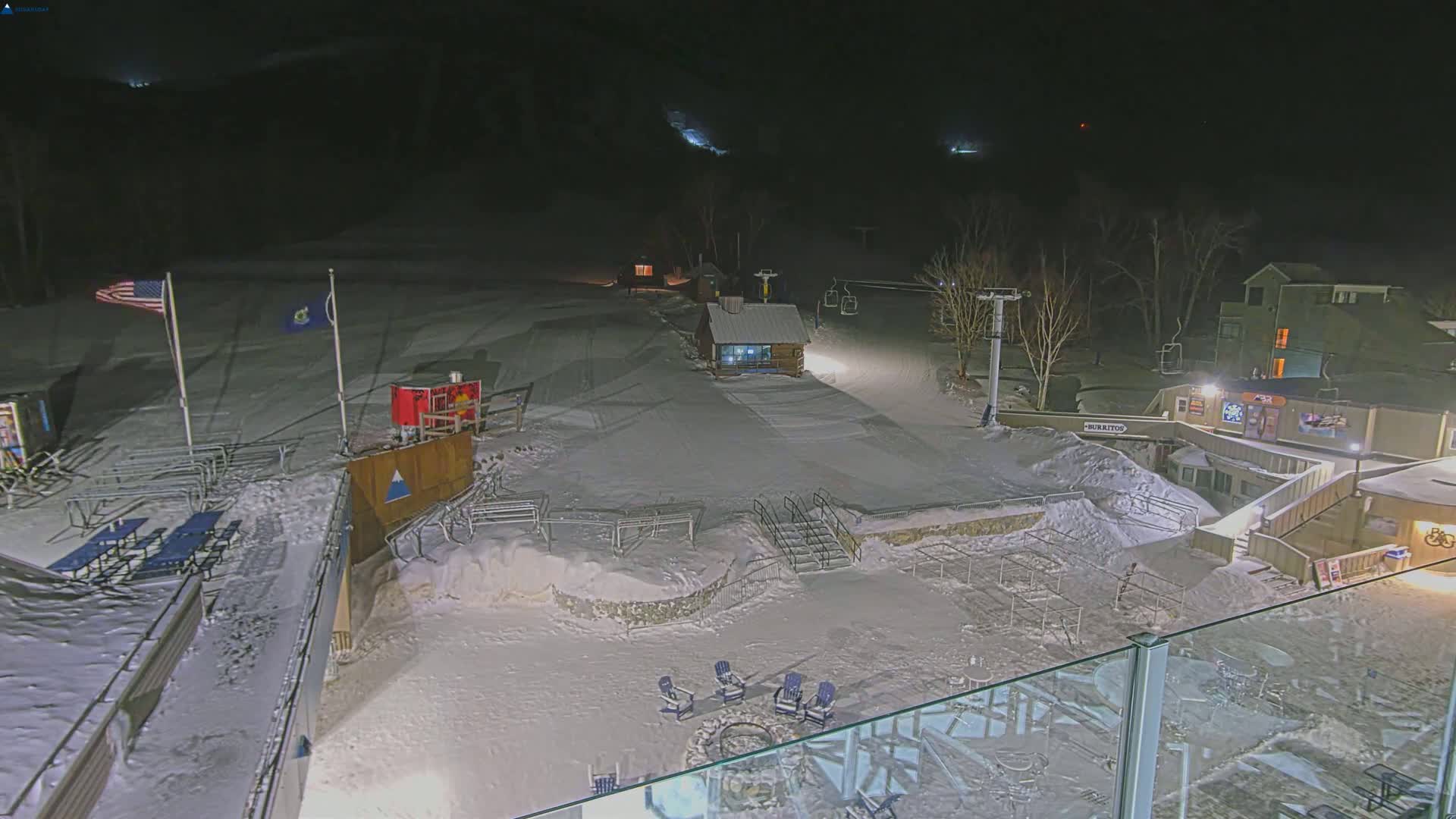 A snow-covered ski resort at night, illuminated by artificial lights, features a small building, ski lift, and outdoor seating areas.