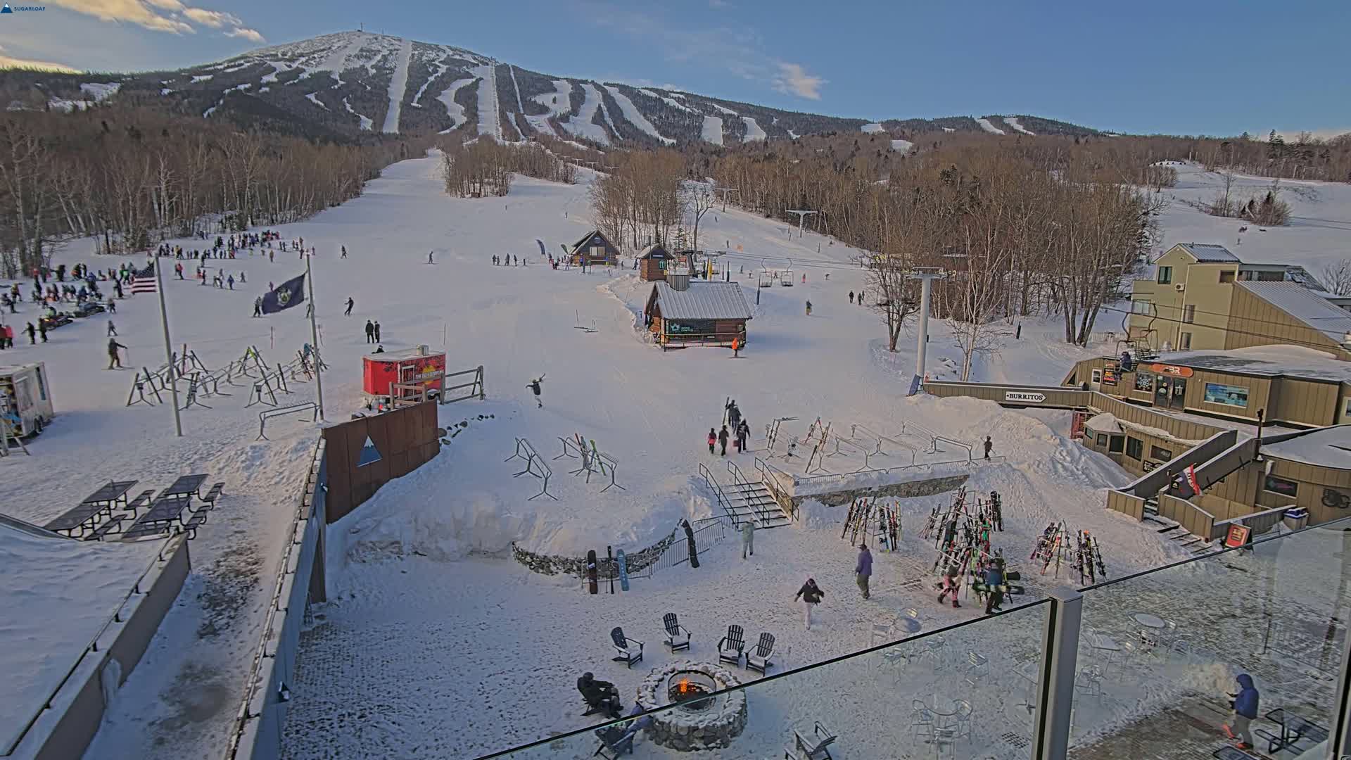A snow-covered ski resort with people skiing and gathering around a fire pit, under a mostly sunny sky, in front of a mountain with ski slopes.
