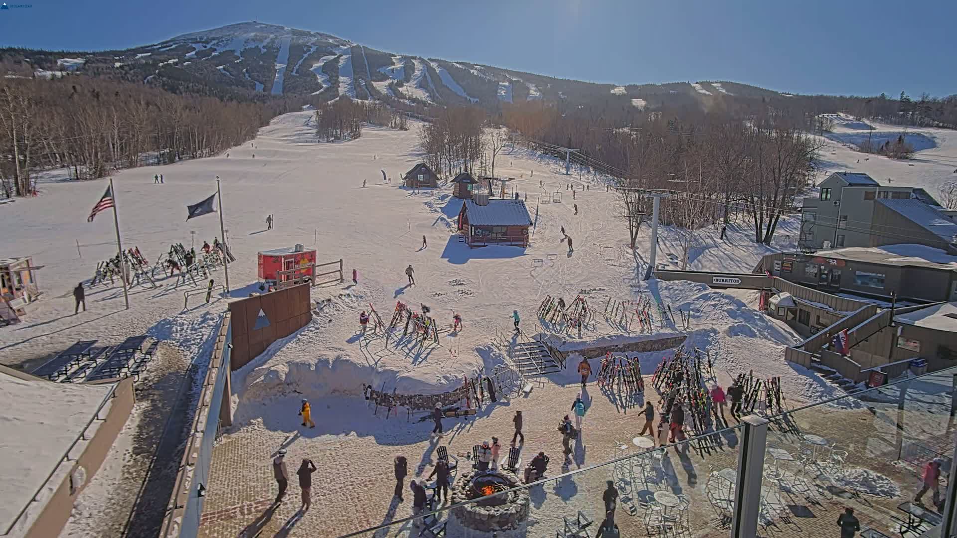 A sunny day at a snow-covered ski resort shows skiers and snowboarders on the slopes, with people gathered around a fire pit at the base.