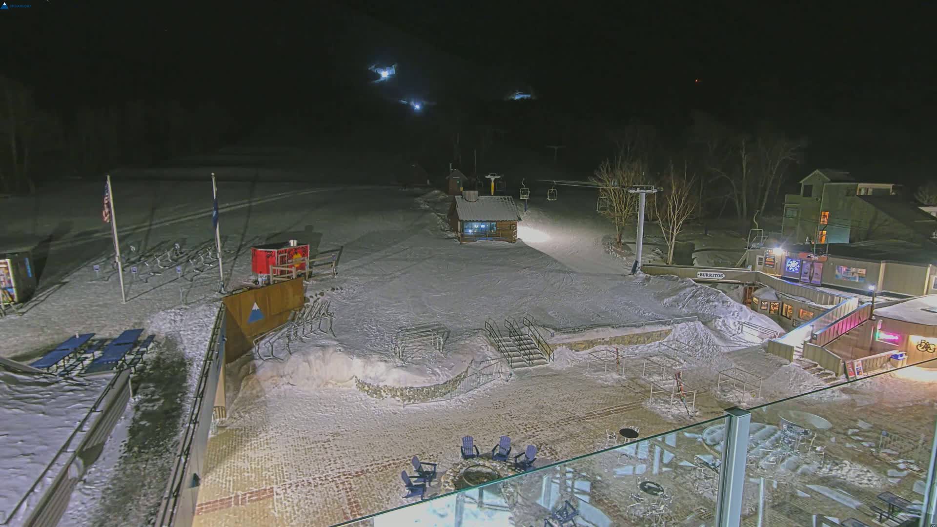 A snow-covered ski resort at night, illuminated by artificial lights, shows a ski lift, small buildings, and outdoor seating areas.