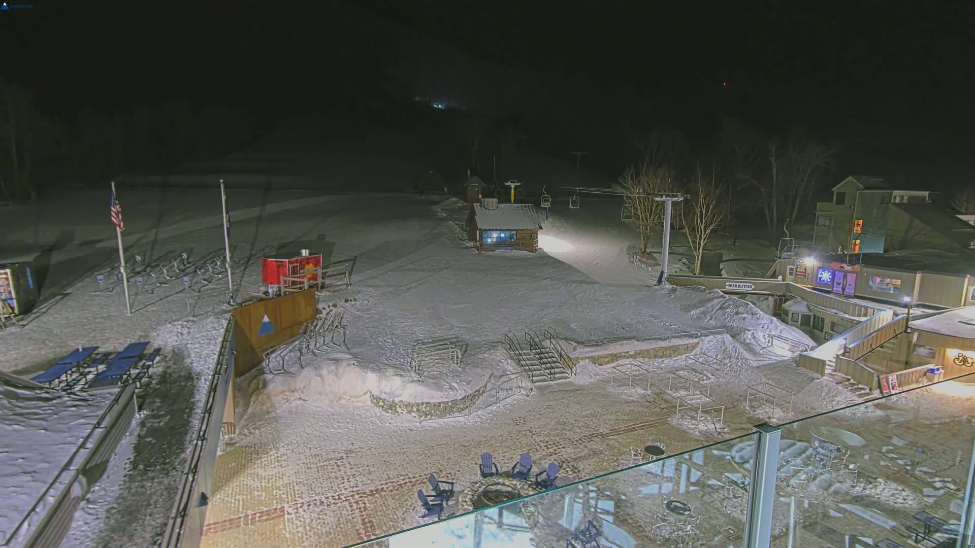 A snow-covered ski resort at night, illuminated by artificial lights, features a ski lift, small buildings, and outdoor seating areas.
