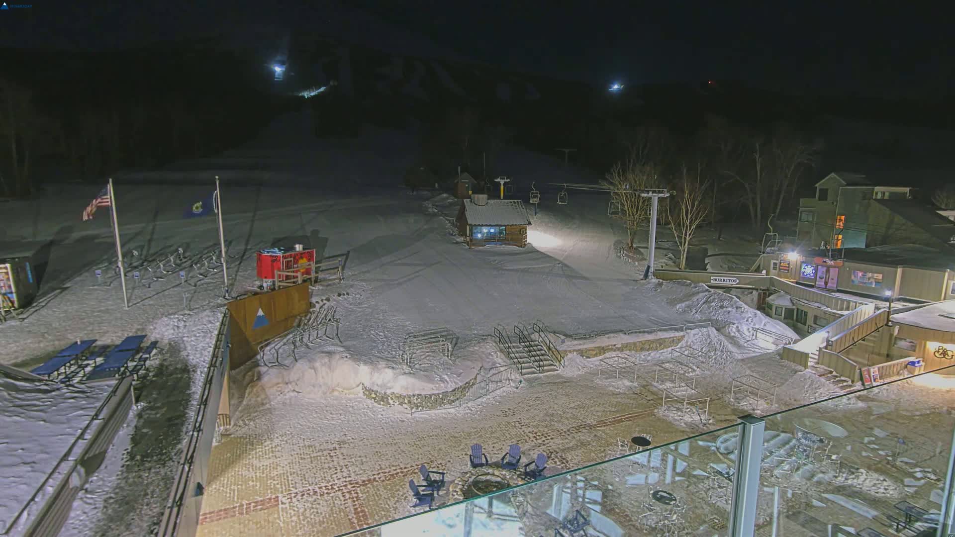 A nighttime view of a snow-covered ski resort base area with a chairlift, small buildings, and outdoor seating areas.