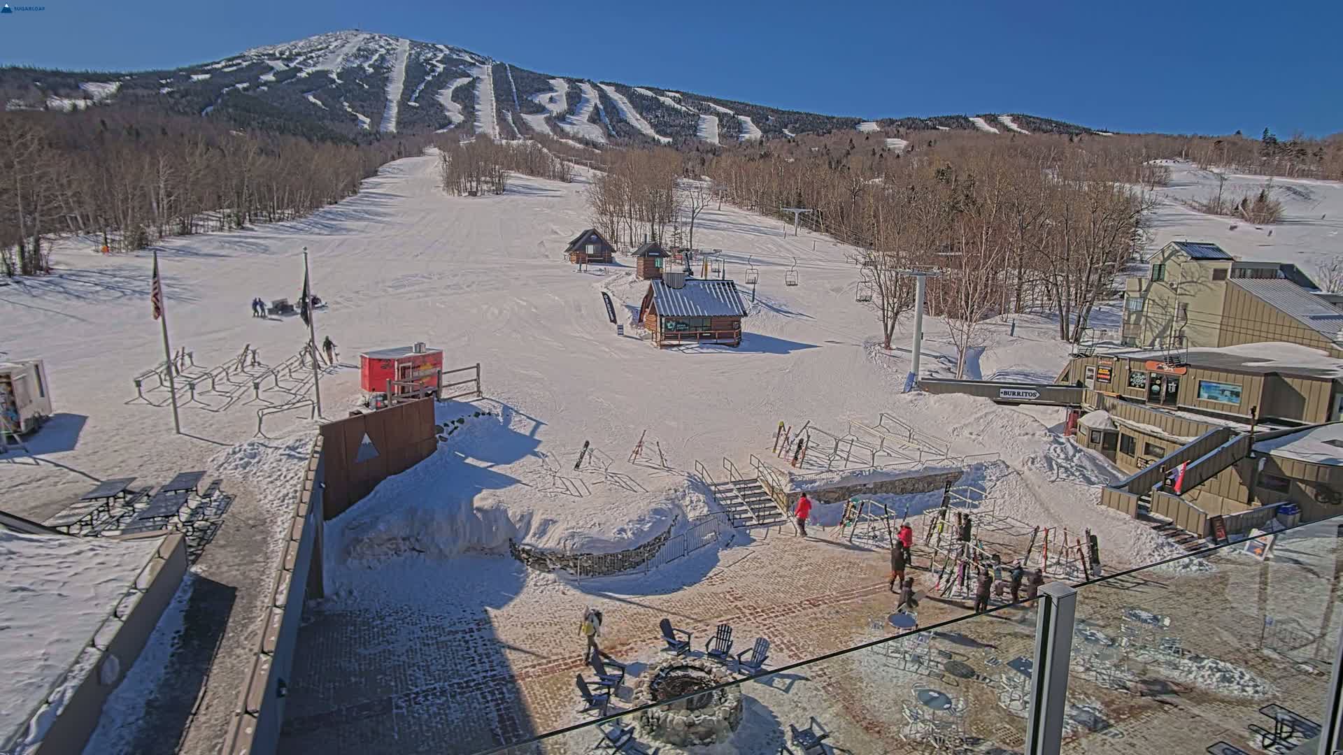 A snow-covered ski resort on a sunny day, with several small buildings, a fire pit, and skiers visible near the base of a large mountain with numerous ski runs.