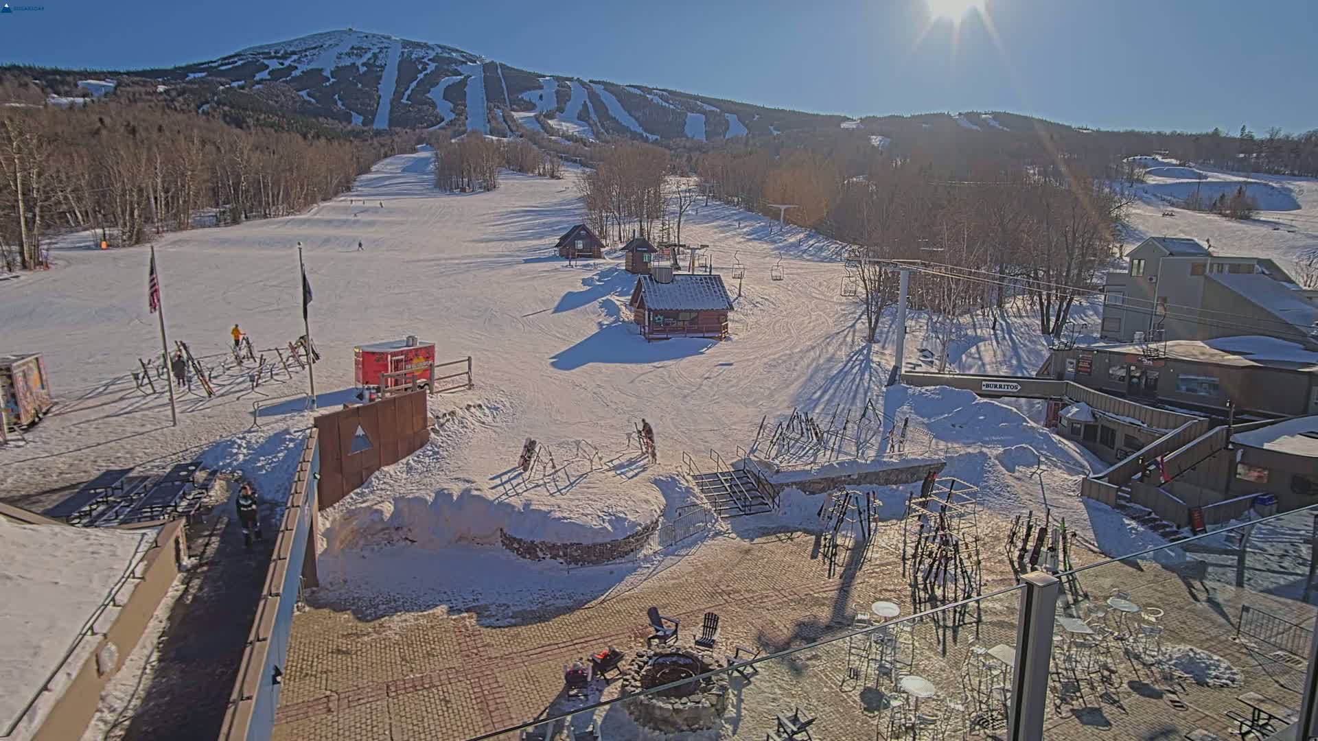 A sunny winter day at a ski resort shows a snow-covered mountainside with ski runs, several small buildings, and outdoor seating areas with a fire pit.